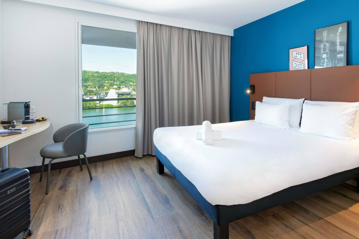 Een hotelkamer met een bed, een bureau, een stoel, een koffer en uitzicht op een brug en groen.