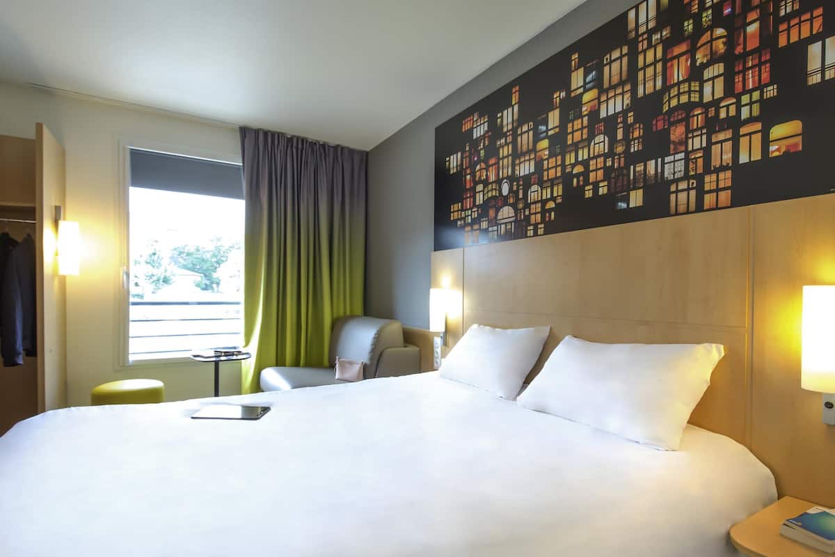 Ein Hotelzimmer mit einem großen Bett, einem Sessel, einem kleinen Tisch und Blick ins Freie.