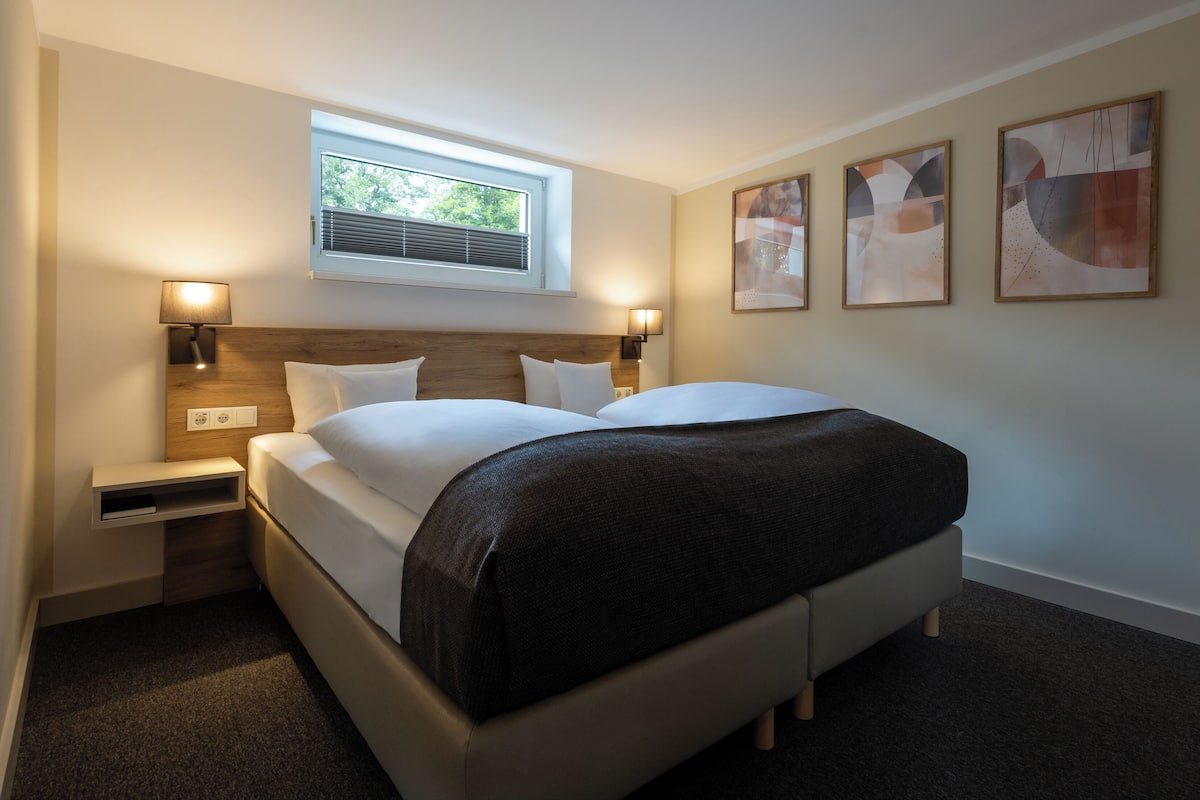 A modern hotel room with a large bed, bedside lamps, a window with blinds, and abstract wall art.