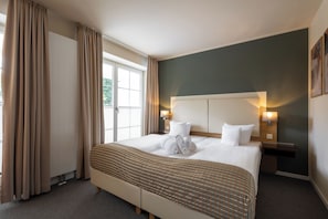 A hotel room with a large bed, beige curtains, a view of greenery outside, and a dark green accent wall.