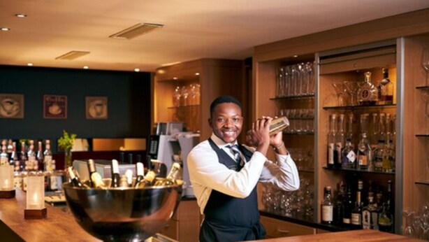 Een bar met een bartender, een grote metalen kom en schappen vol flessen en glazen.