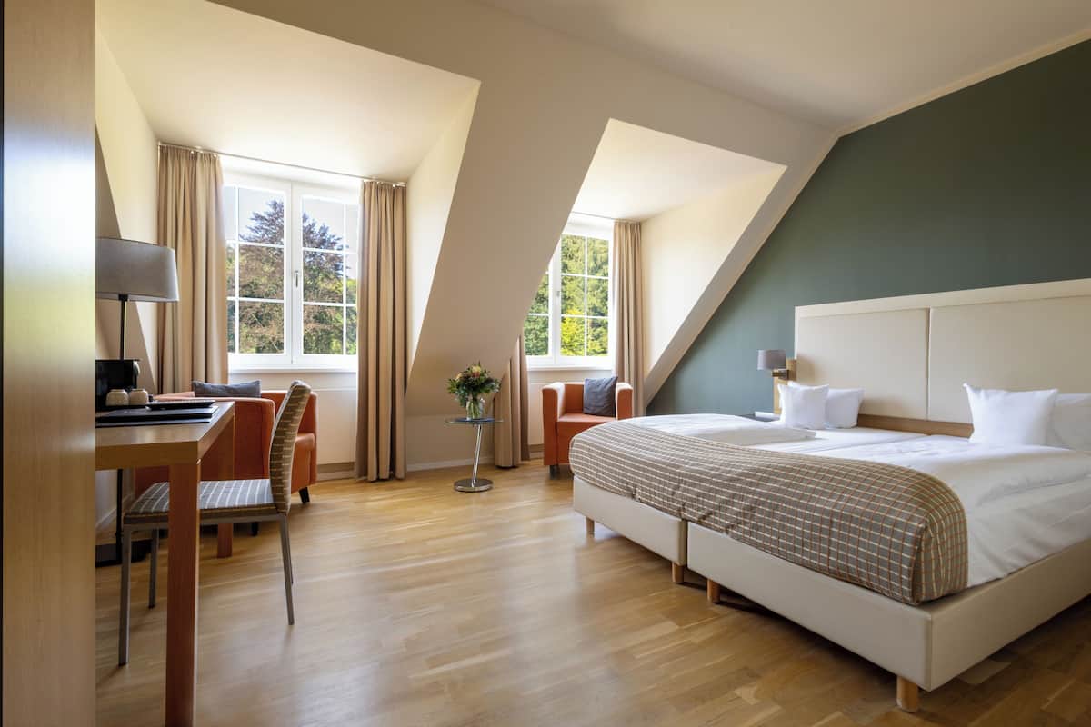 A modern hotel room with a large bed, a desk with a chair, and a view of trees through the windows.