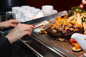 A person slicing a large, cooked meat dish with mushrooms and vegetables on a metal tray.