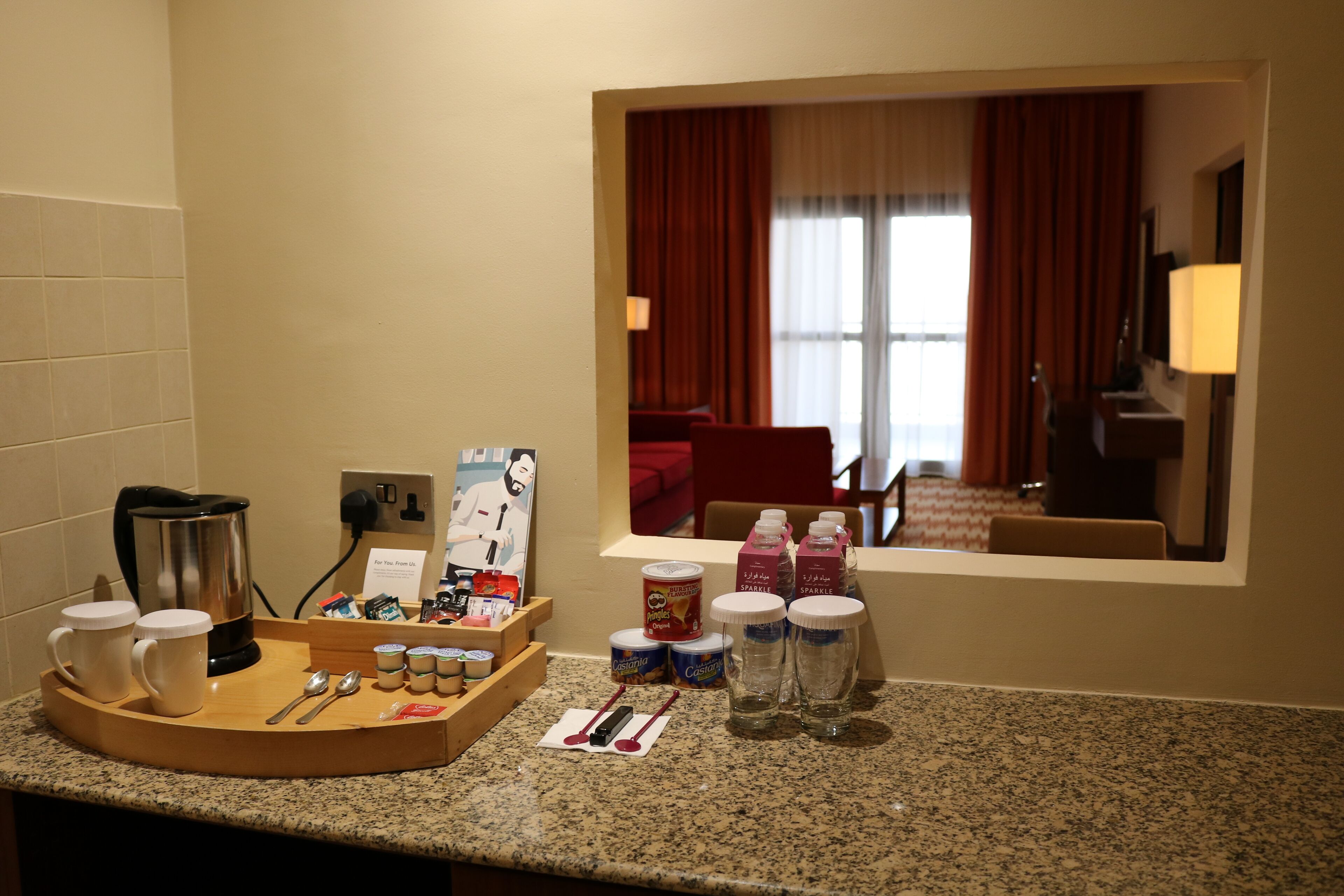 A hotel room's breakfast area with a coffee maker, mugs, a tray with utensils, and a window view of the room.