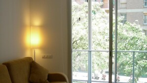 A room with a sofa, a lamp, and a sliding glass door leading to an outdoor area with trees and a building in the background. - Travessera Parc Guell Apartamentos (Barcelona)