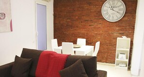 A living room with a brick wall, a large clock, white furniture, and a red throw on the sofa. - Travessera Parc Guell Apartamentos (Barcelona)