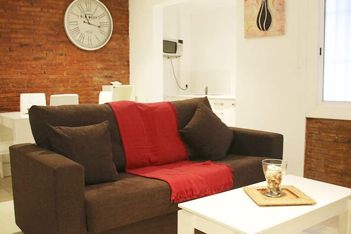 A living room with a brown sofa, white coffee table, and a red throw blanket. A clock is visible on the wall, and there is a window with blinds.