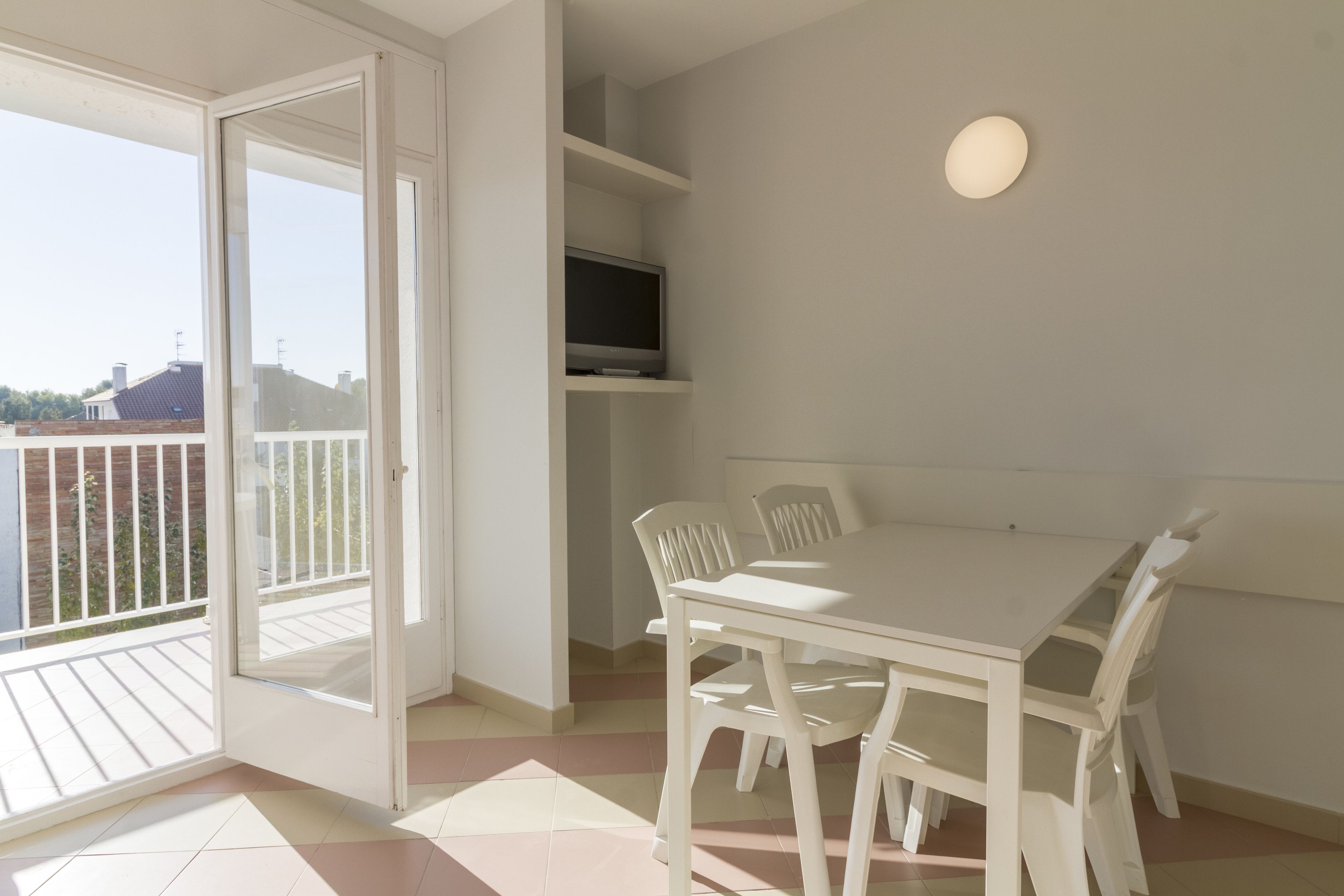 A room with a white table and chairs, a flat-screen TV, and a sliding glass door leading to an outdoor balcony.