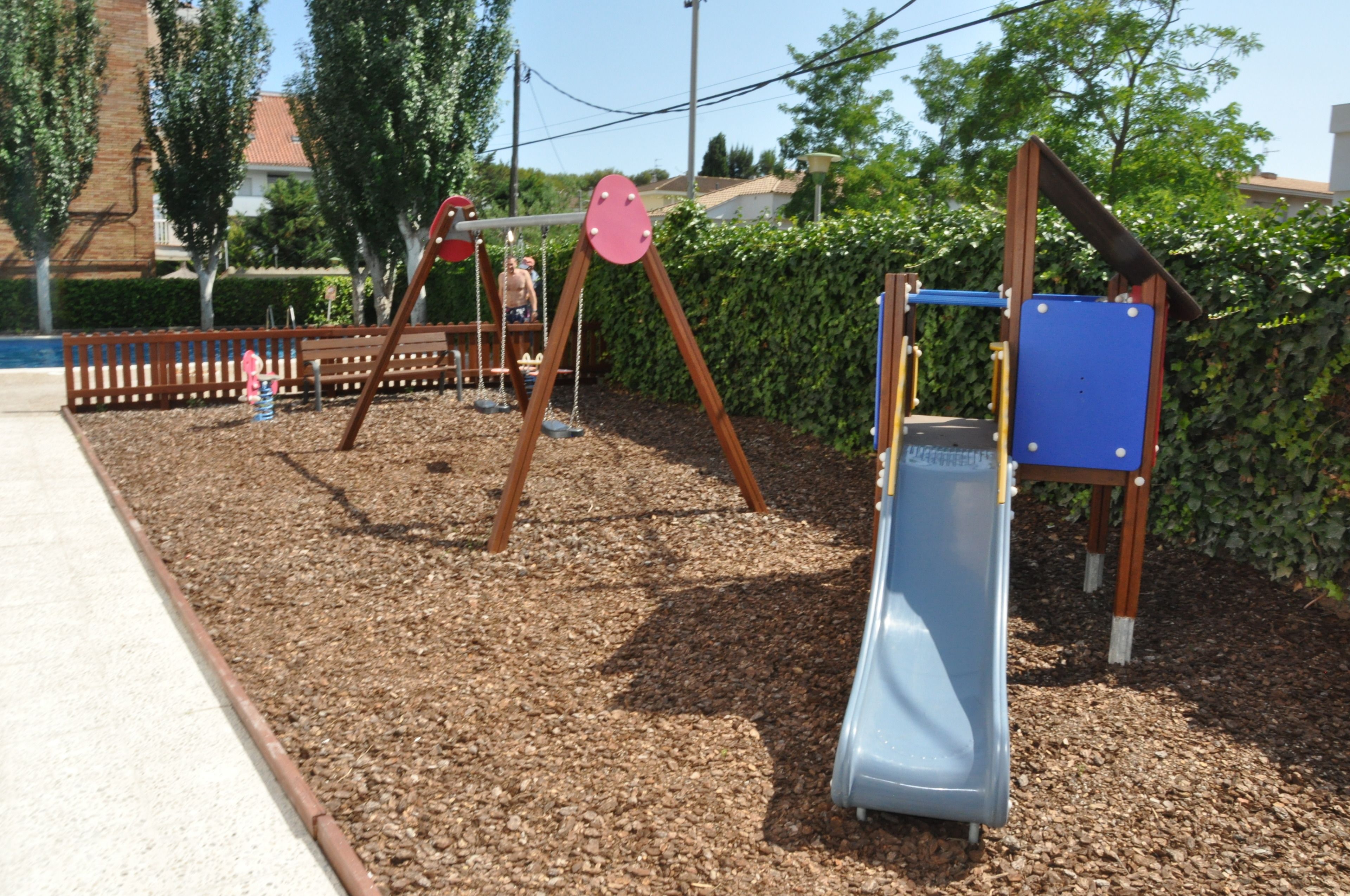 Une aire de jeux avec des balançoires et un toboggan, entourée de paillis et d’arbres.
