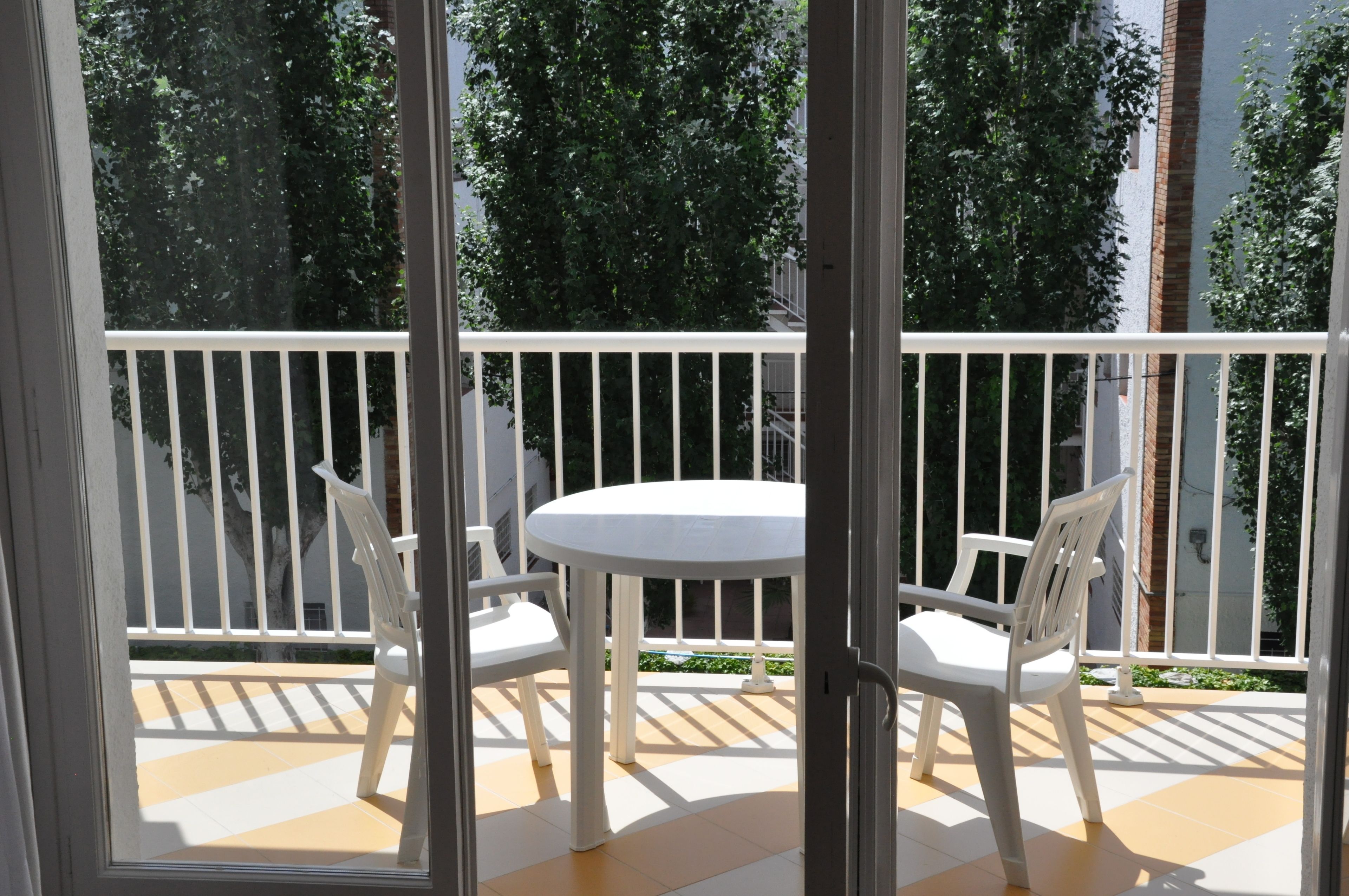 A balcony with a white table and chairs, a white railing, and a view of trees and buildings.
