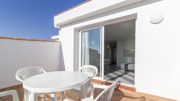 Un patio exterior con piso de baldosas, una mesa redonda y sillas, y un ventanal corredizo que da a una habitación interior.