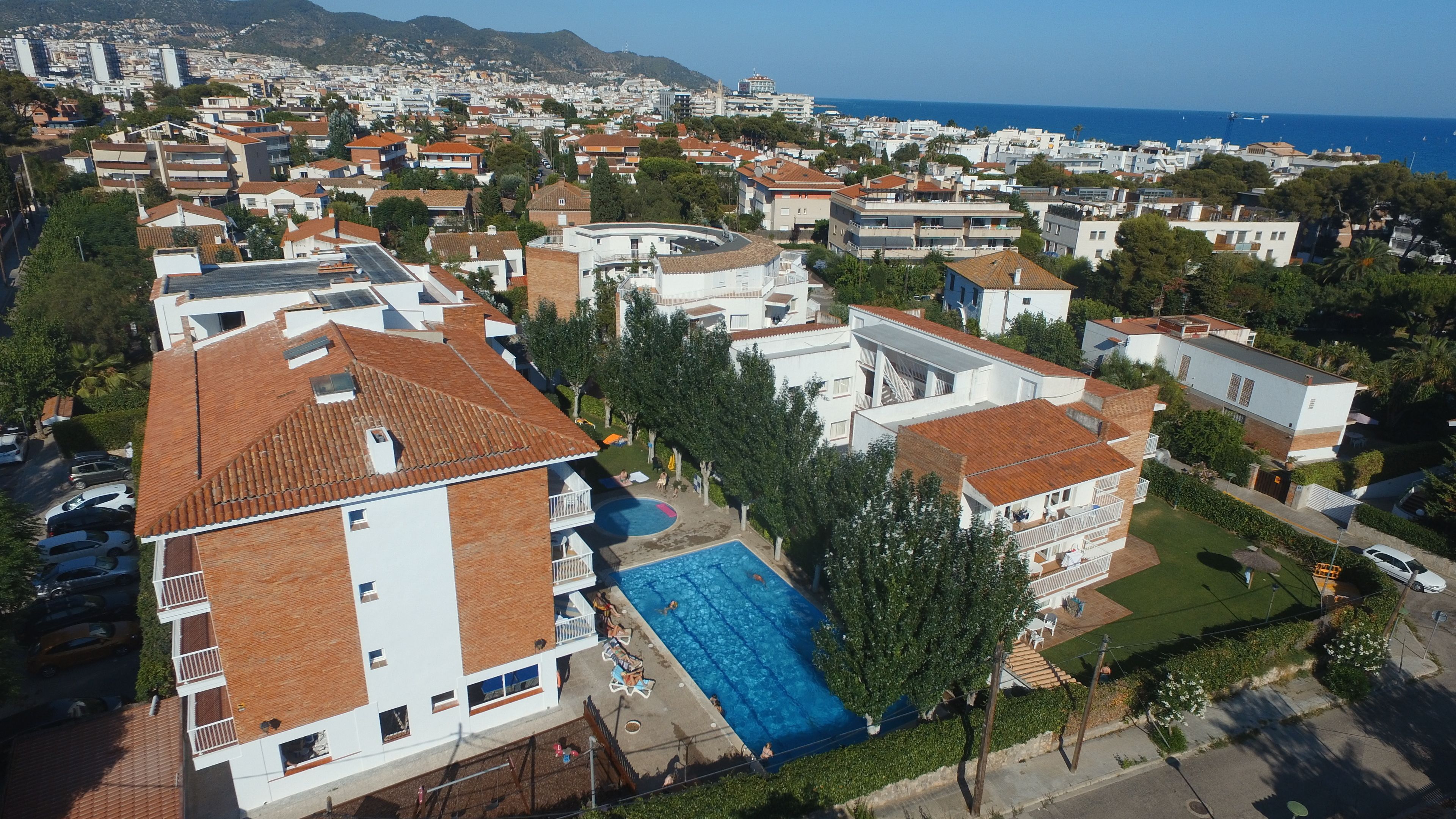 A residential area with a swimming pool surrounded by houses, a clear view of the sea, and a hilly landscape in the background.