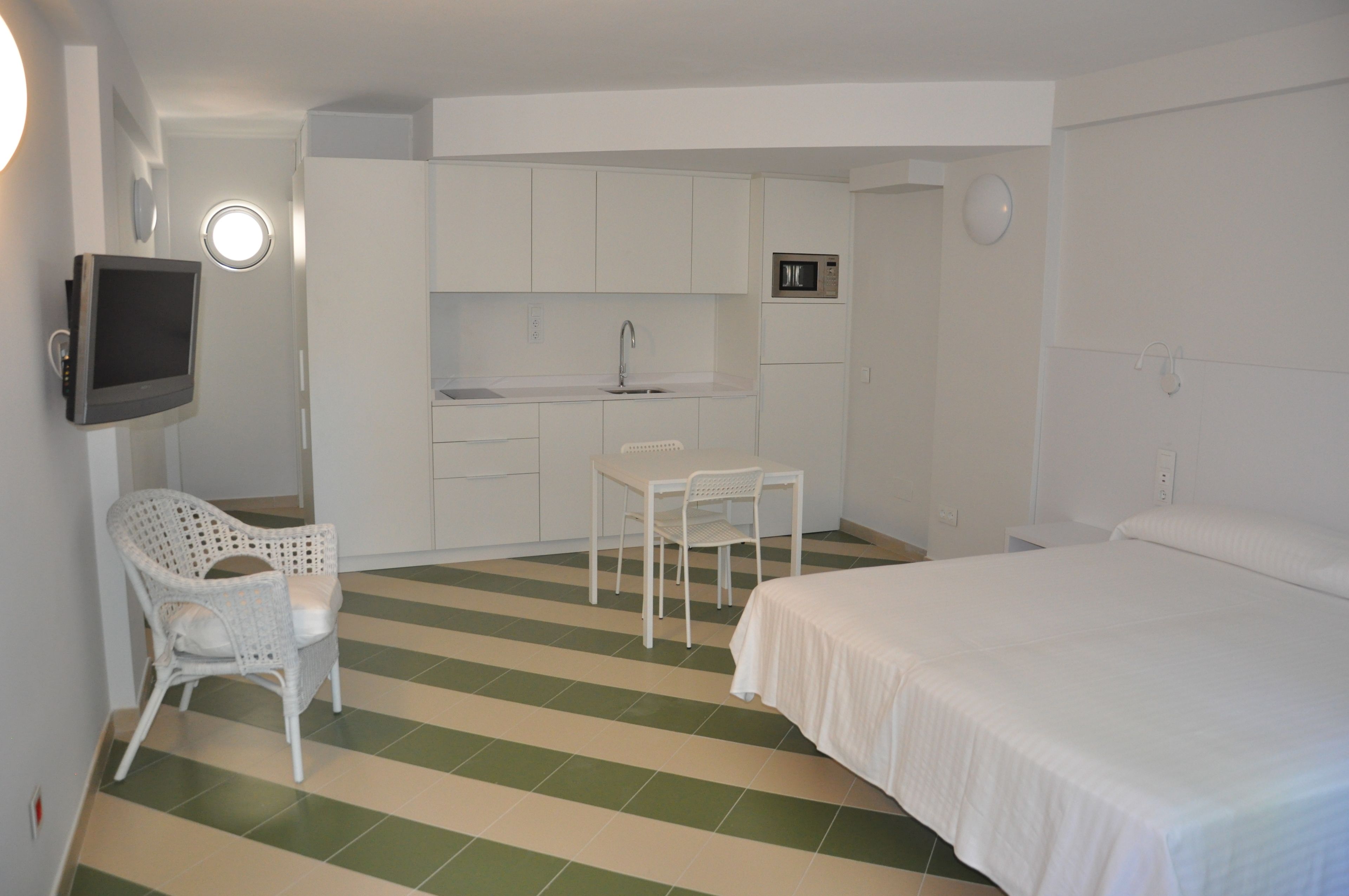 A compact hotel room with a bed, a small kitchenette, a TV, and a chair.