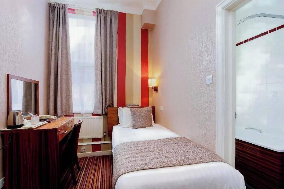 Habitación de hotel con cama, escritorio, silla, ventana con cortinas, lámpara de pared y puerta.
