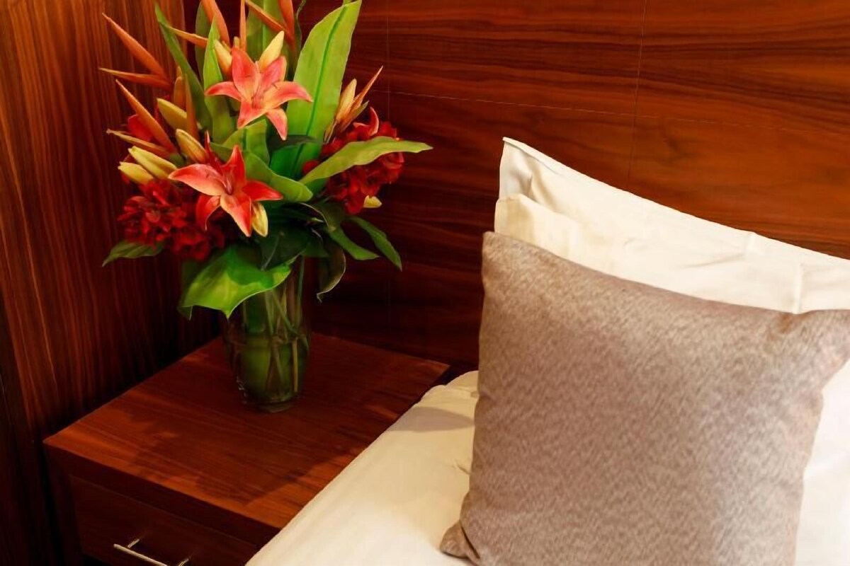 A wooden nightstand with a vase of flowers and a pillow on a bed.
