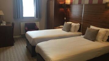 A hotel room with two beds, a wardrobe, a desk with a lamp, and a window with curtains.