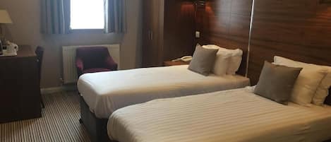A hotel room with two beds, a wardrobe, a desk with a lamp, and a window with curtains.
