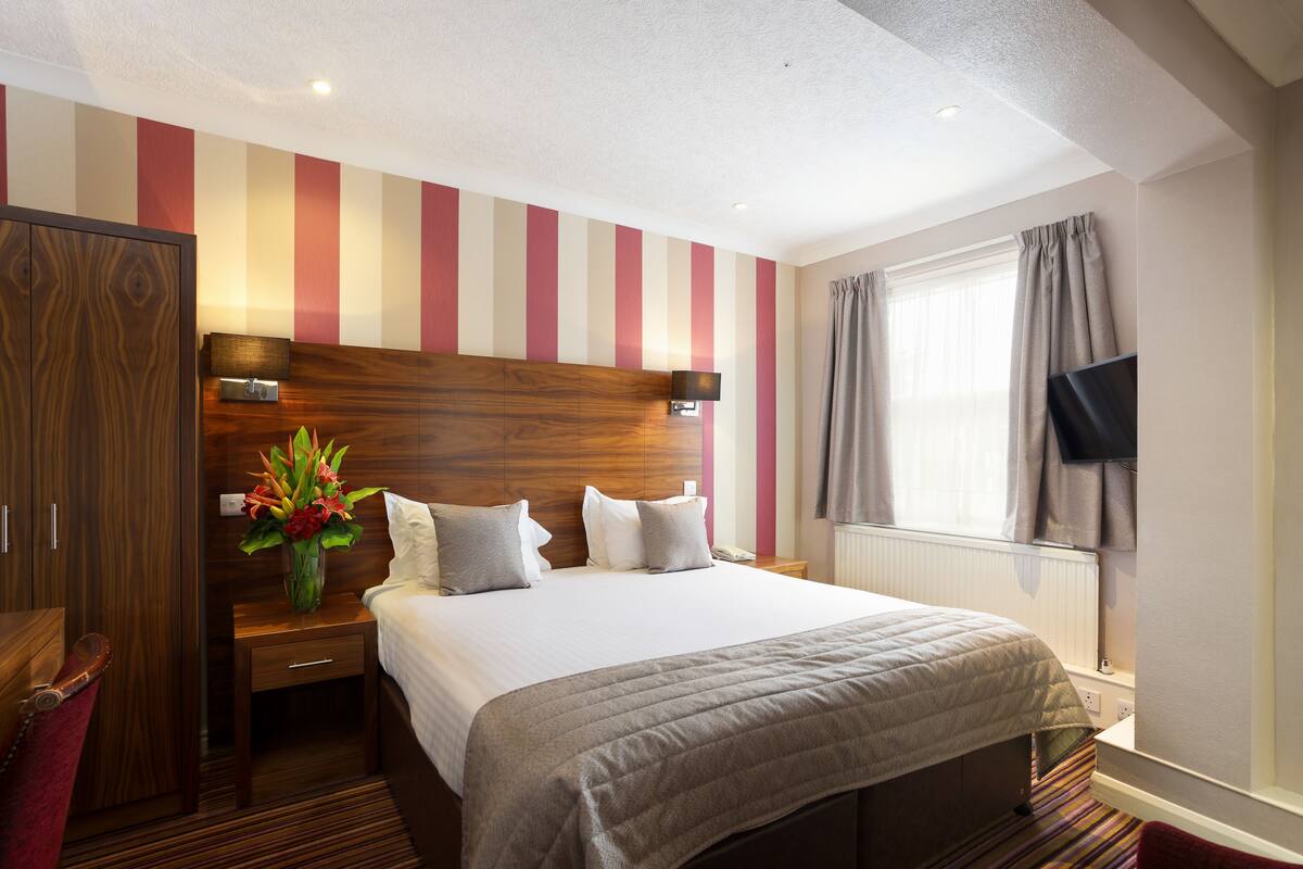Habitación de hotel con una cama grande, cabecera de madera, mesita de noche con lámpara, televisor montado en la pared y una ventana con cortinas.