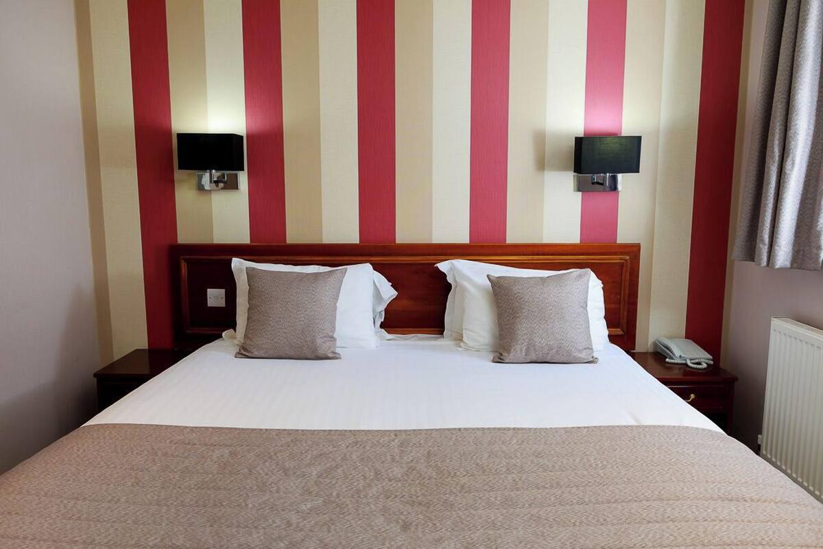 Habitación de hotel con pared a rayas, cabecera de madera, dos mesitas de noche, cama con sábanas blancas y dos almohadas, y televisor montado en la pared.