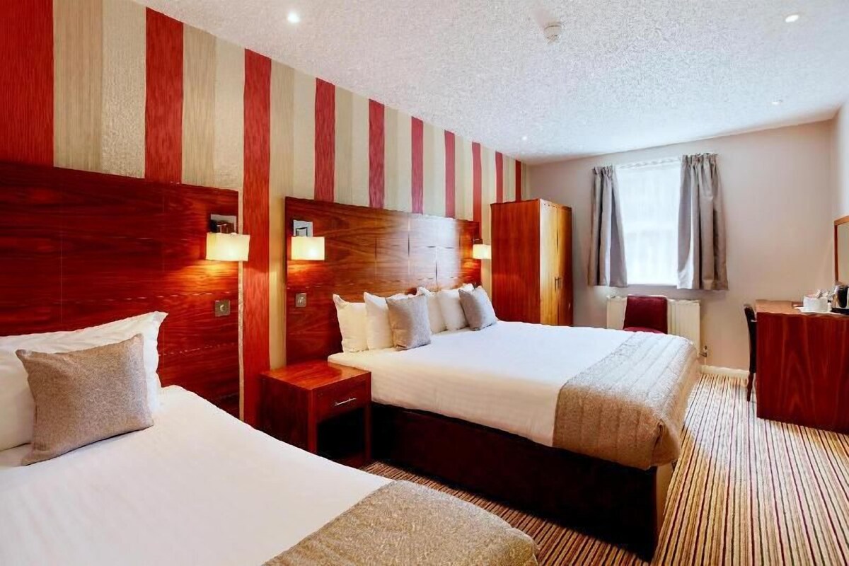 A hotel room with two beds, a wooden headboard, bedside lamps, a window with curtains, and a desk with a chair.