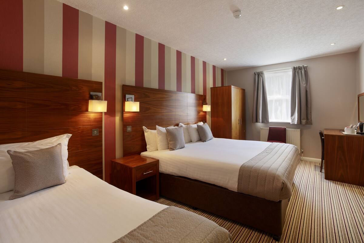 Una habitación de hotel con dos camas, cabeceros de madera, lámparas de noche, un escritorio y una ventana con cortinas.