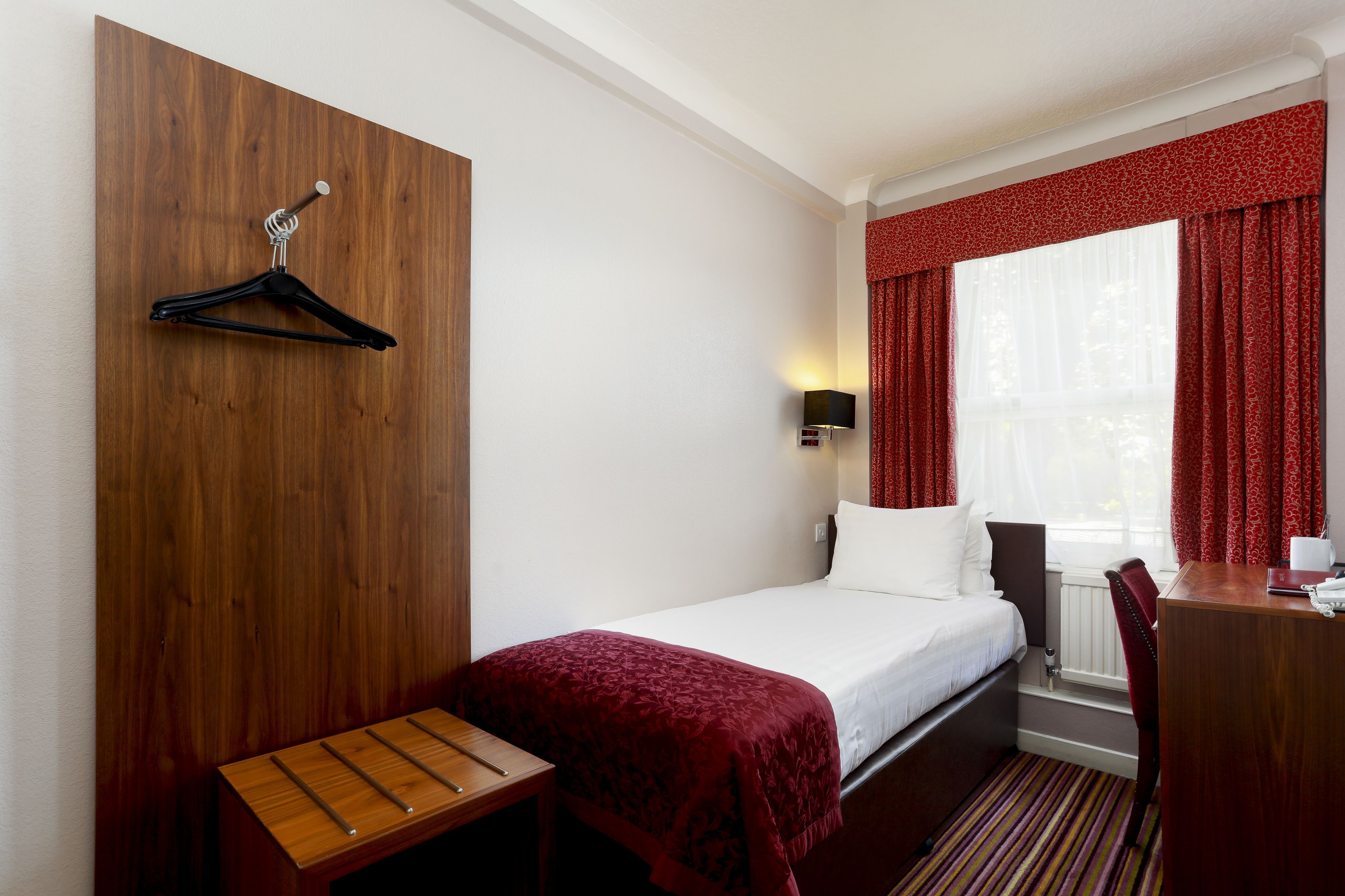 A hotel room with a bed, a wooden desk, a chair, a TV, and a window with red curtains.