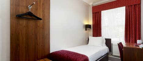 A hotel room with a bed, a wooden desk, a chair, a TV, and a window with red curtains.