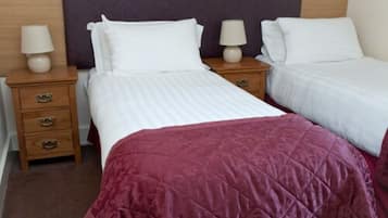 Quarto de hotel com duas camas de solteiro, cada uma com roupa de cama branca e uma mesa de cabeceira de madeira com luminária.