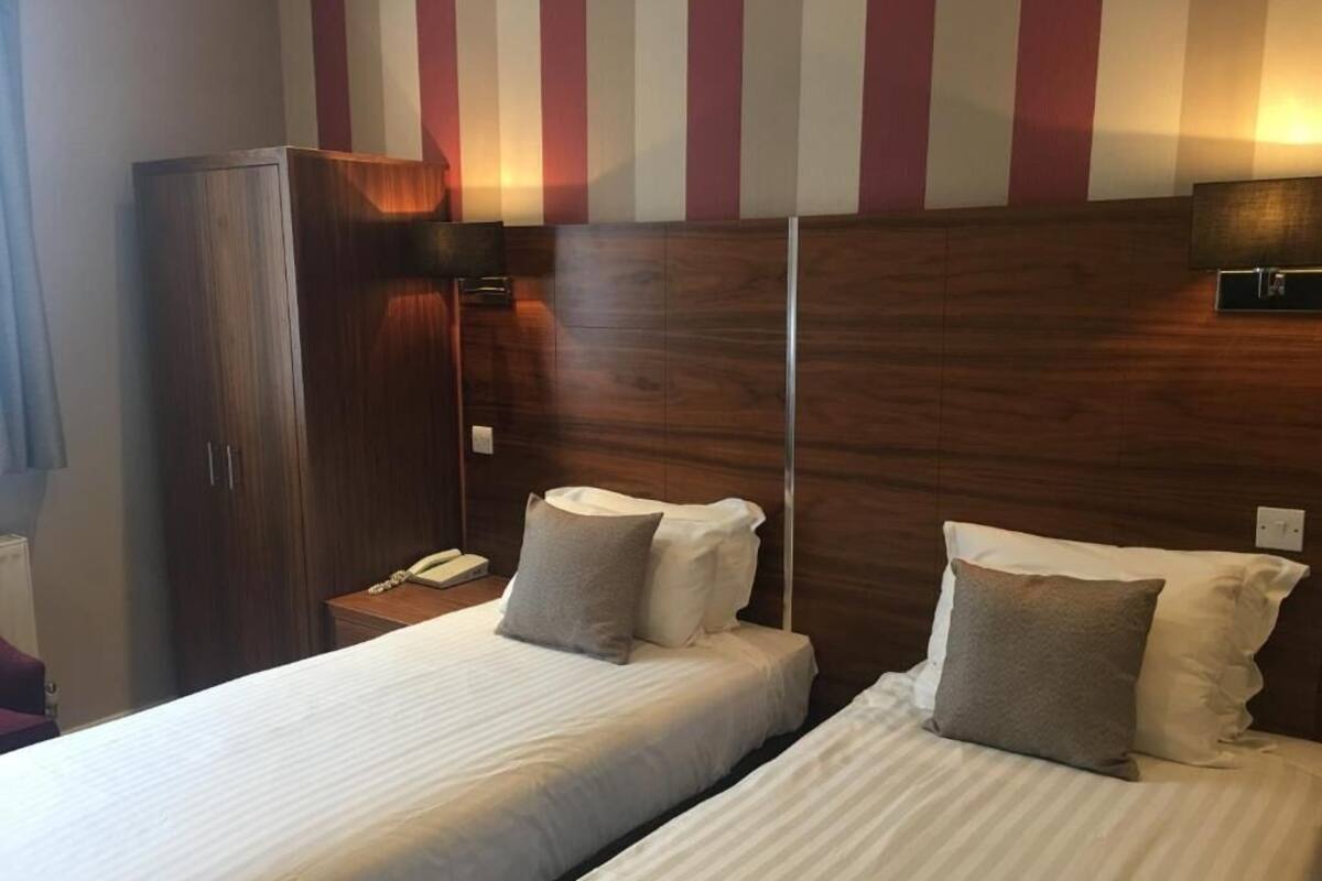Habitación de hotel con dos camas, cabecero de madera, un armario y una mesita de noche con lámpara.