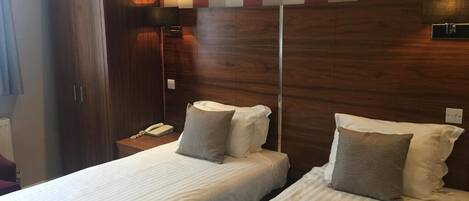 A hotel room with two beds, wooden headboard, a wardrobe, and a bedside table with a lamp.