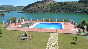 Una piscina con un camino de piedra que la conecta, rodeada por una cerca y con vistas a un lago y colinas.
