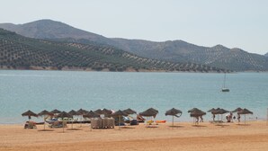 Una playa con sombrillas de paja y sillas de playa, un bote y un velero en el agua.