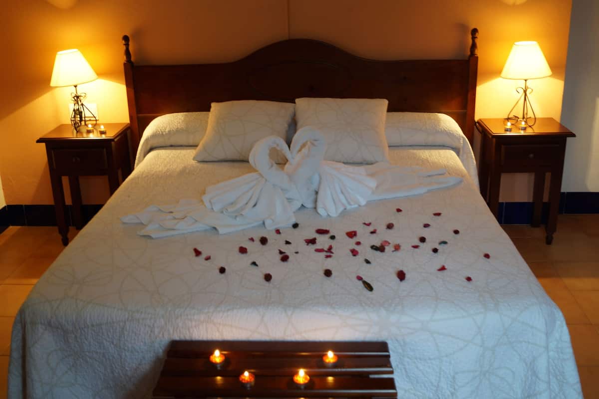 Una cama bien hecha con edredón blanco, dos almohadas y una toalla dispuesta en forma de cisne. Hay pétalos de rosa esparcidos sobre la cama y una bandeja con velas al pie de la cama.