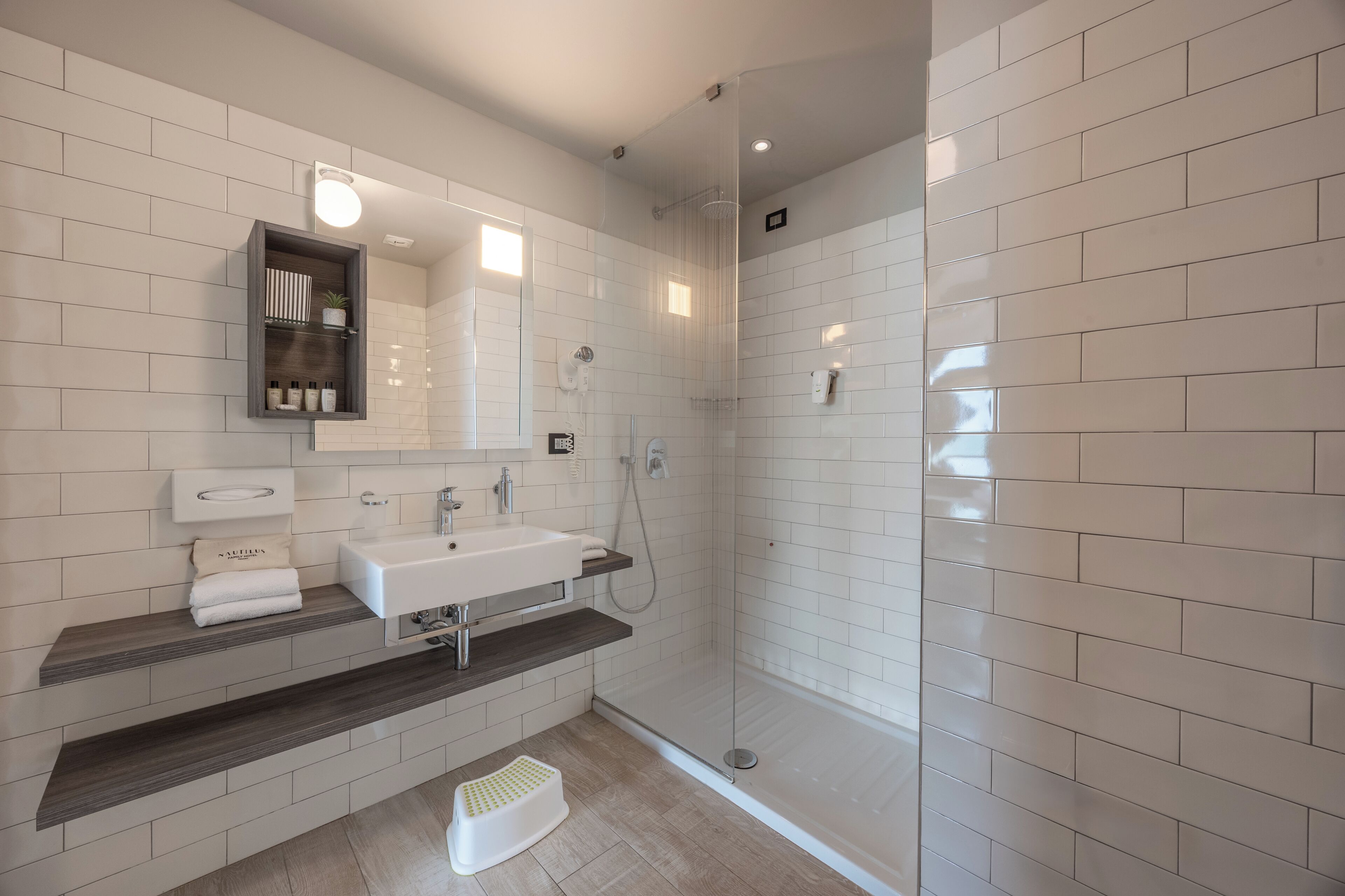 Un baño moderno con ducha de azulejos blancos, un lavabo con espejo y un estante con artículos de aseo.
