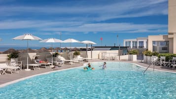 Piscina com espreguiçadeiras e guarda-sóis, rodeada por edifícios e um céu claro.