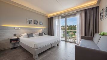 A modern hotel room with a large bed, a sofa, a nightstand with a lamp, and a view of the outdoors.