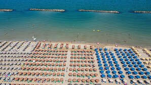 A beach with rows of sun umbrellas and lounge chairs, a clear blue sea, and a rocky breakwater. - Nautilus Family Hotel (Pesaro)