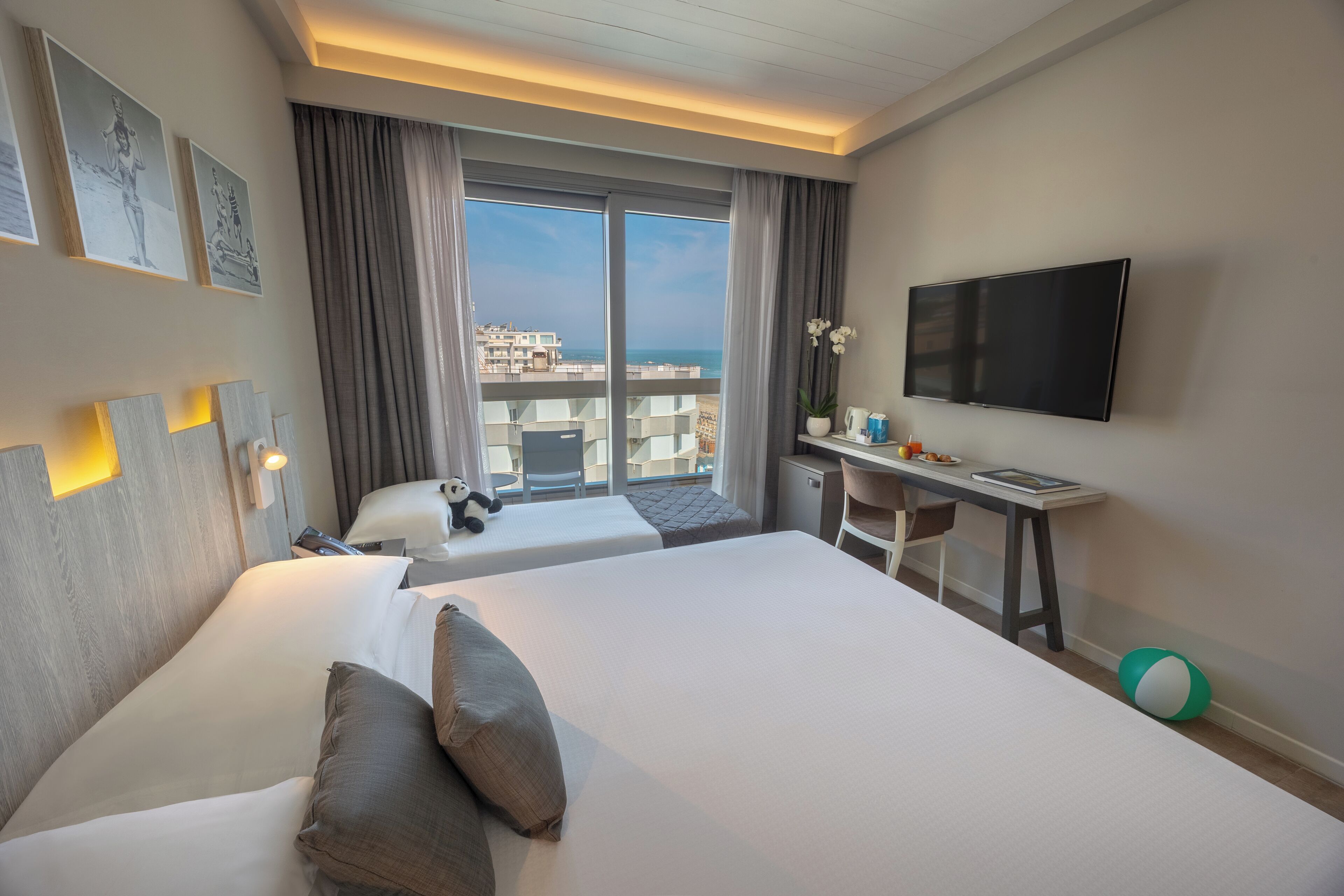 Ein Hotelzimmer mit zwei Betten, einem Schreibtisch, einem Fernseher und einem Balkon mit Blick auf die Stadt.