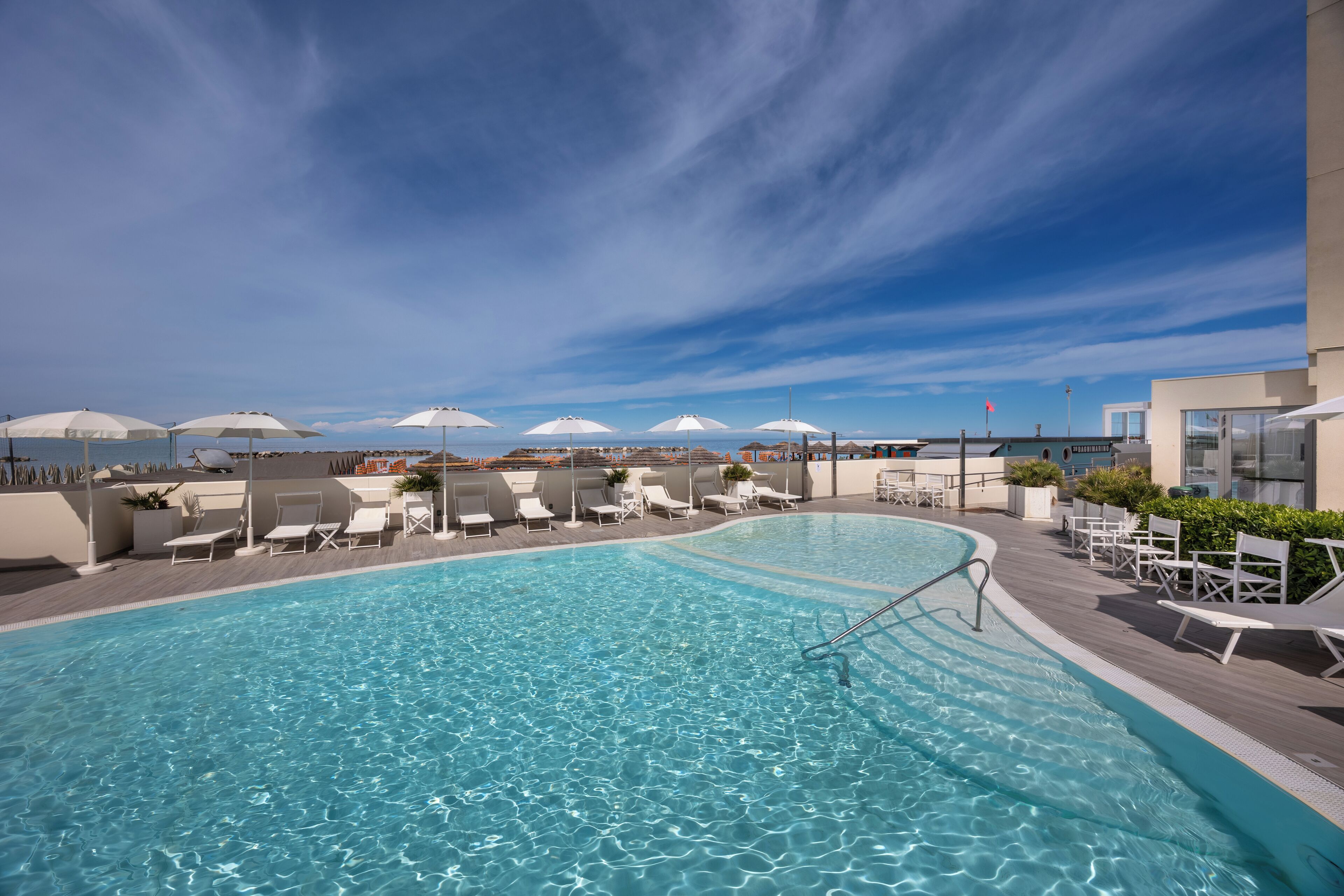 A rooftop pool with clear blue water, surrounded by white lounge chairs and umbrellas, overlooking a cityscape.