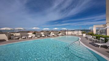 A rooftop pool with clear blue water, surrounded by white lounge chairs and umbrellas, overlooking a cityscape.