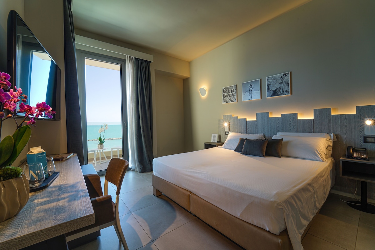 A hotel room with a large bed, a desk with a chair, a TV, and a view of the ocean.