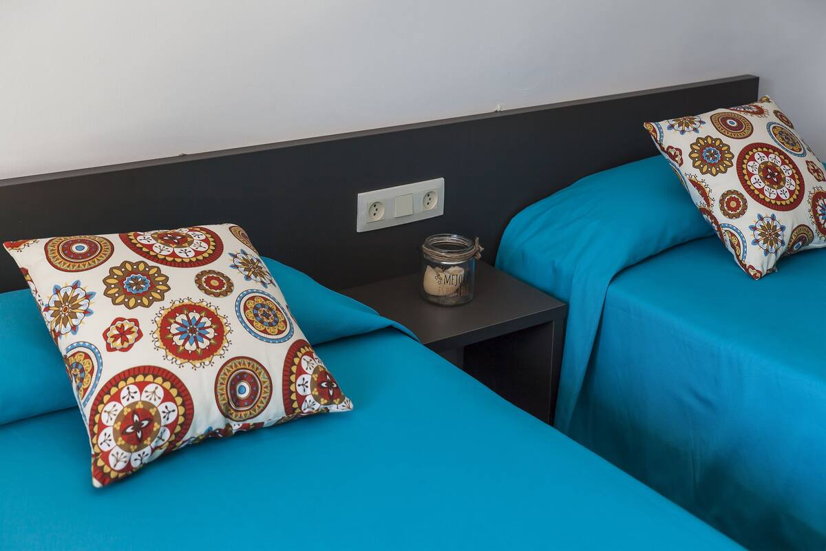 A hotel room with two beds, each with a blue bedspread and a patterned pillow. There is a small table between the beds with a jar on it.