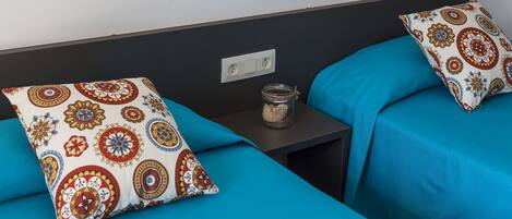 Habitación de hotel con dos camas, cada una con una colcha azul y un cojín estampado. Una mesita pequeña entre las camas sostiene un tarro.