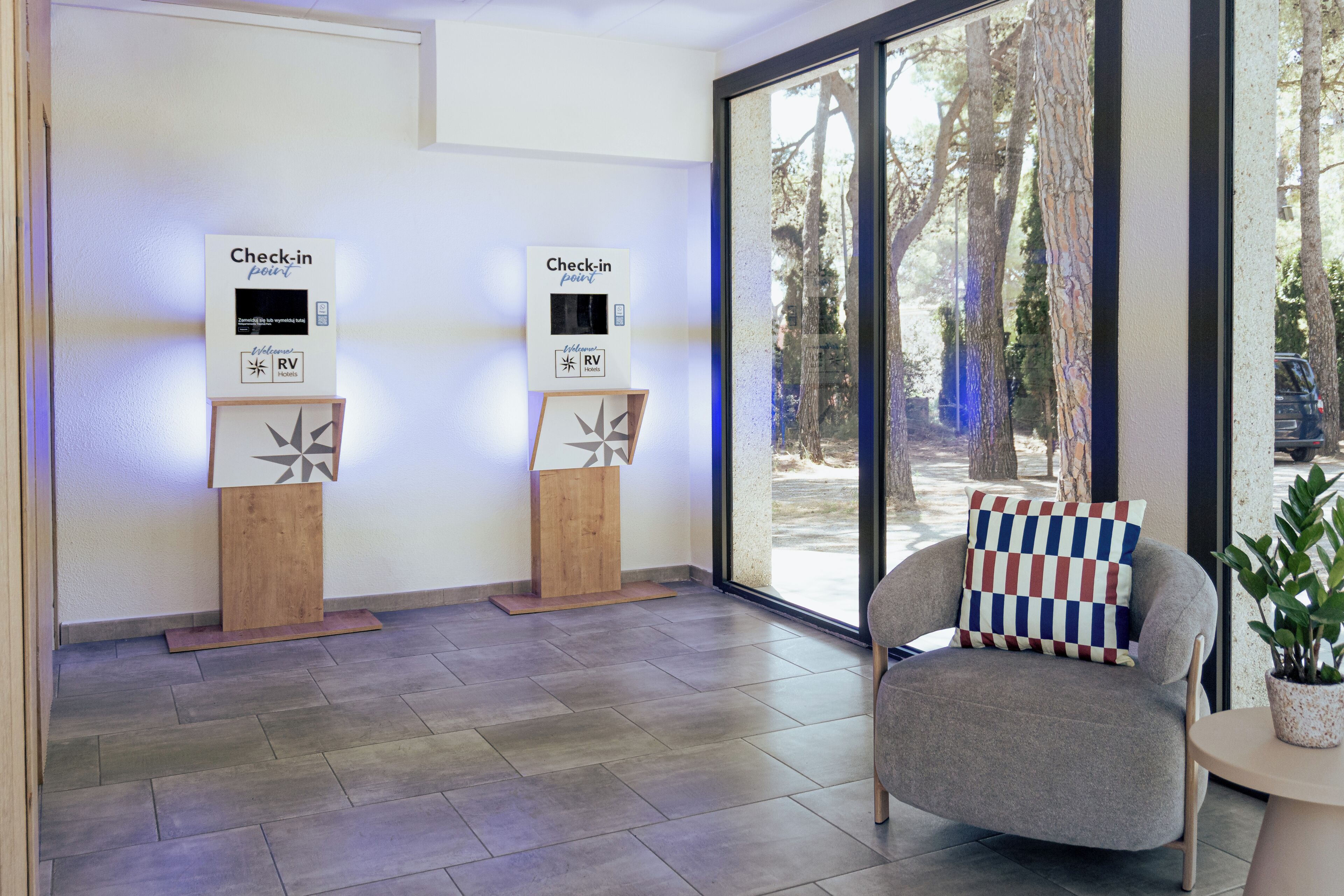 Un punto di check-in con due chioschi self-service, una poltrona grigia con cuscino a quadretti e vista sugli alberi attraverso un'ampia finestra.