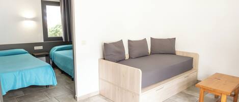 Una habitación pequeña y moderna con sofá cama, dos camas individuales, una mesa de madera y ventana con cortinas.