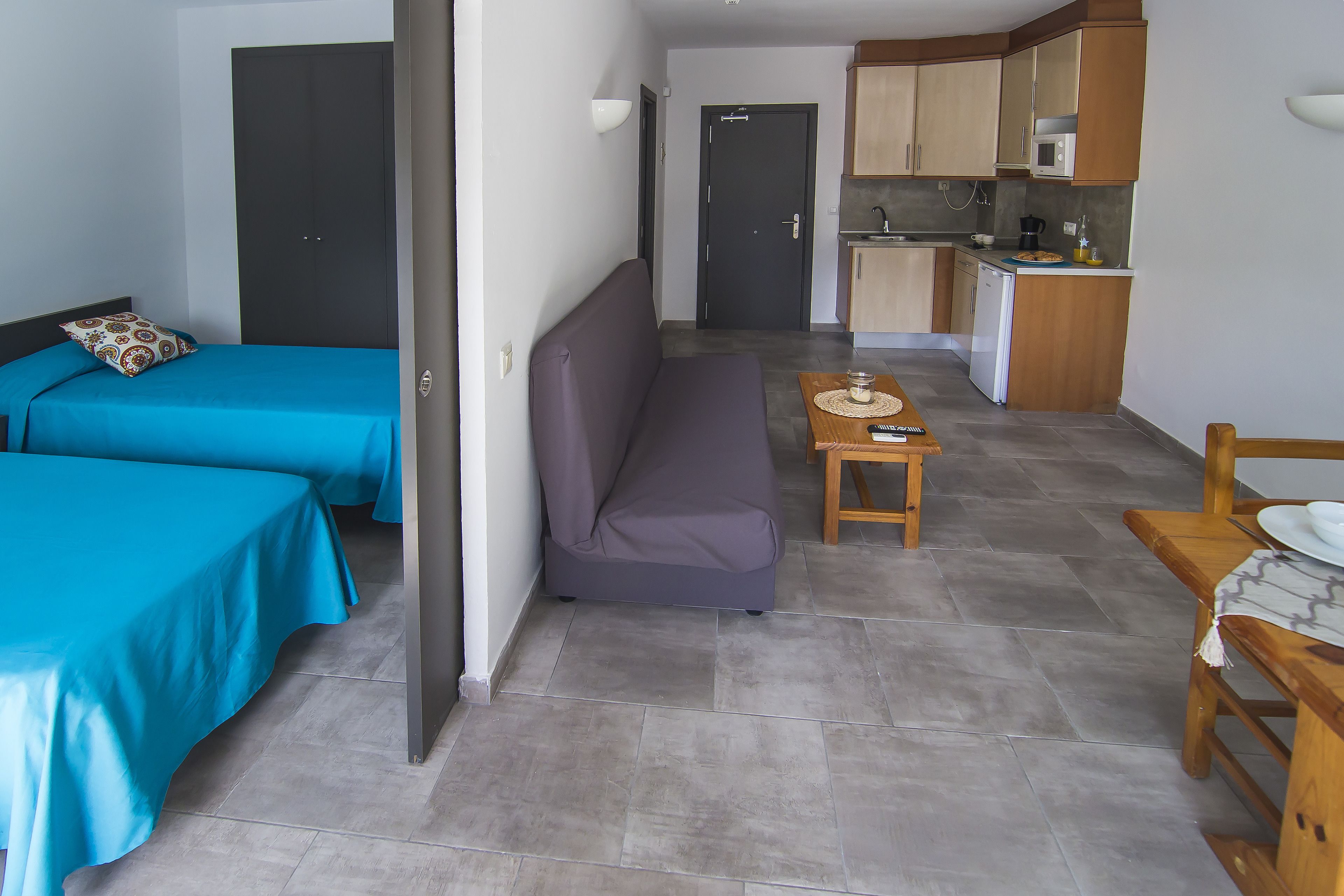 A room with two beds, a sofa, a small table with a cup, and a kitchenette.