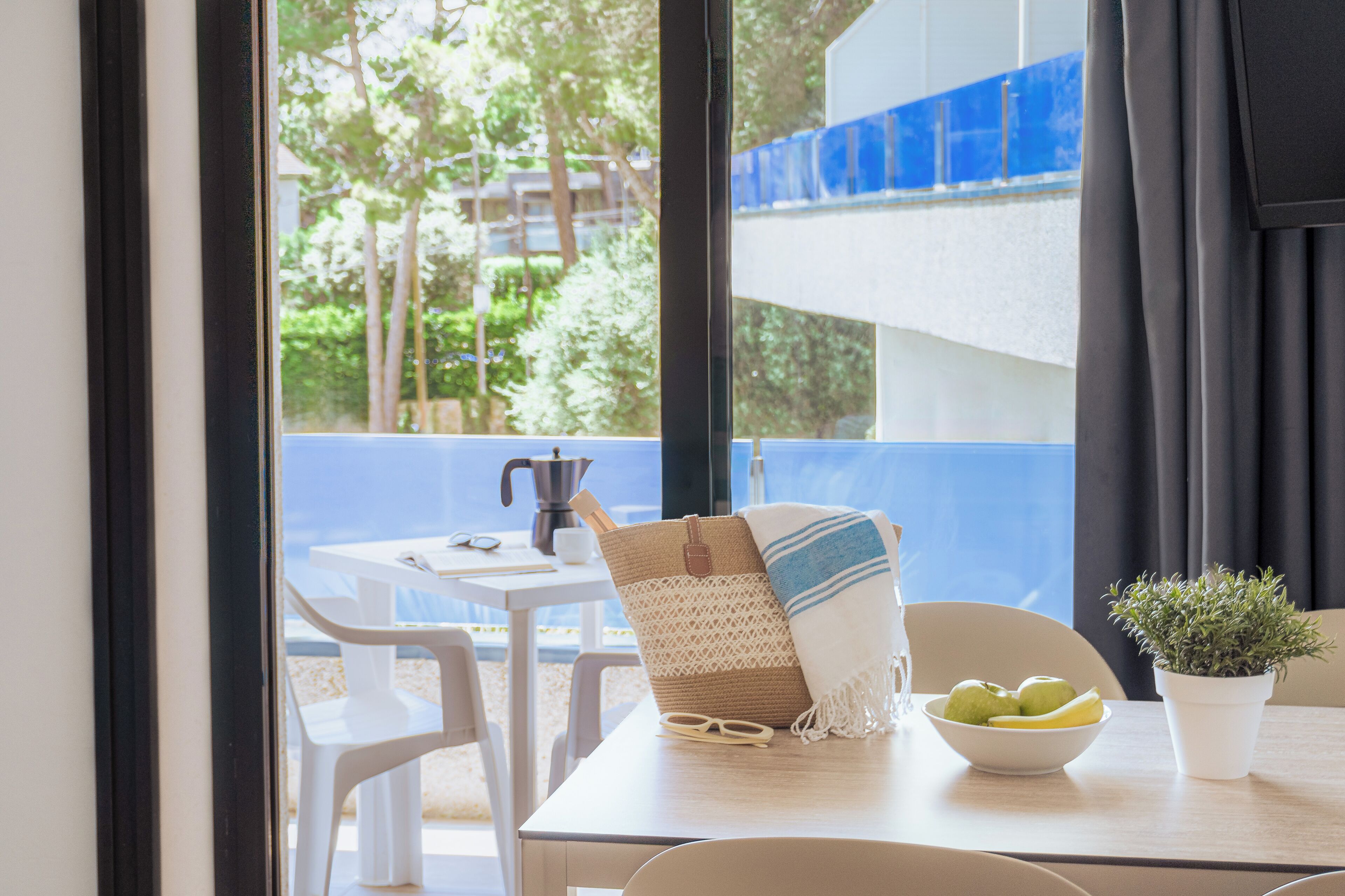 Uma mesa com toalha branca, uma cadeira de vime, uma tigela de frutas, um vaso de planta e uma janela com vista para vegetação e uma estrutura azul.