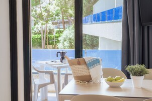A table with a white tablecloth, a woven chair, a bowl of fruit, a potted plant, and a window with a view of greenery and a blue structure.