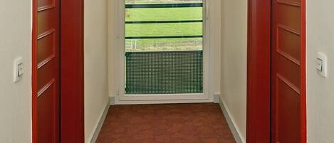 A narrow hallway with red doors, a view of a green field and mountains through a window, and a light fixture on the wall.
