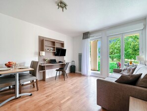 Un salon moderne comprenant un coin repas, un téléviseur à écran plat fixé sur une étagère en bois, un canapé et une grande fenêtre donnant sur un paysage verdoyant.